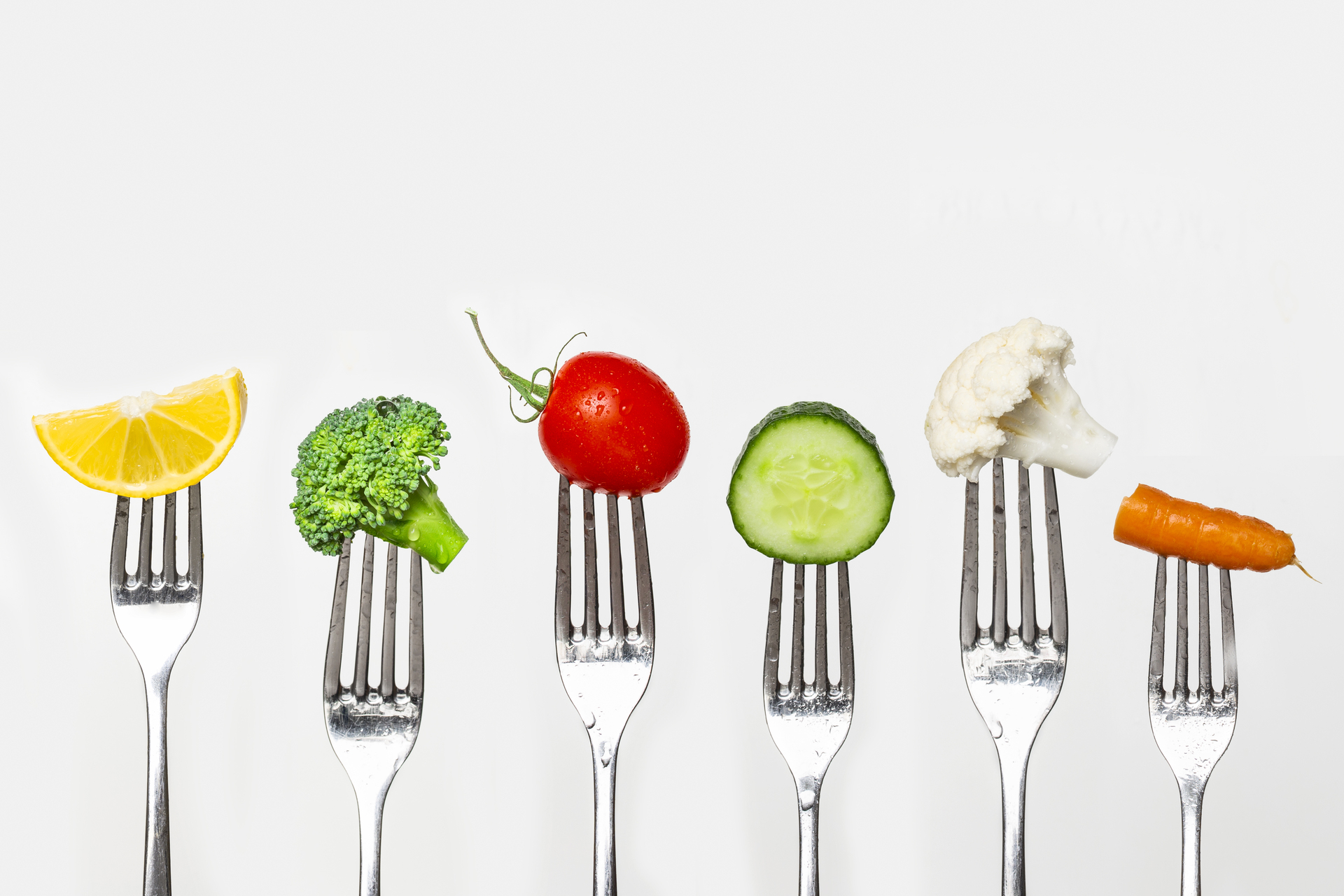 A row of six forks, each holding a piece of fresh vegetable or fruit: a lemon slice, broccoli floret, cherry tomato, cucumber slice, cauliflower floret, and a baby carrot, all against a plain white background.