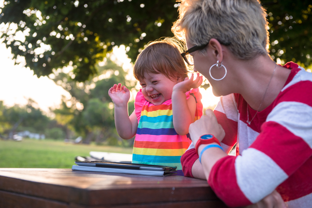 A mother and her young daughter sitting outdoors, smiling and interacting joyfully, with a tablet computer placed on the table in front of them.