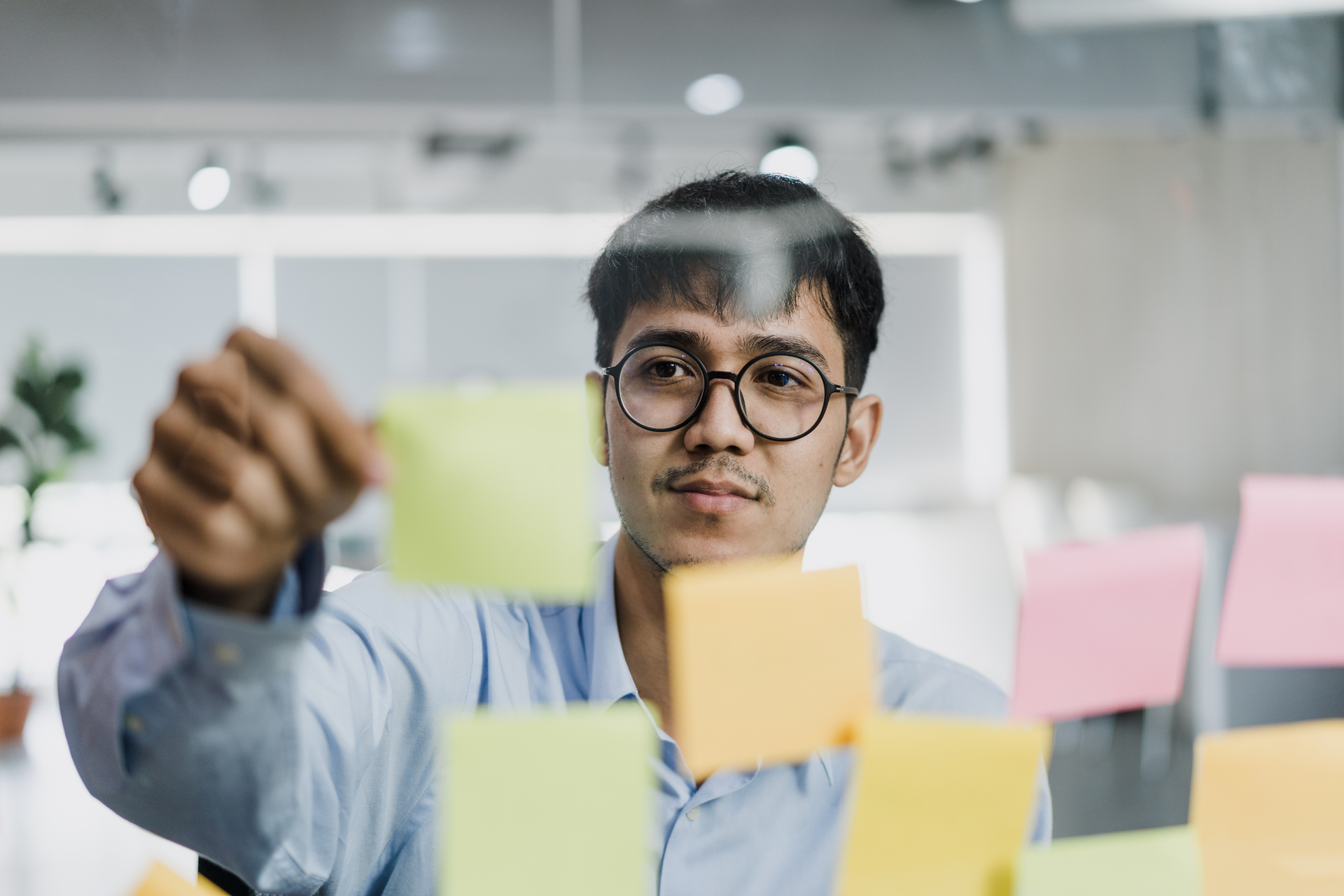 Focused man placing sticky notes on a glass board, symbolizing task management, organisation challenges, or cognitive planning in a work setting.