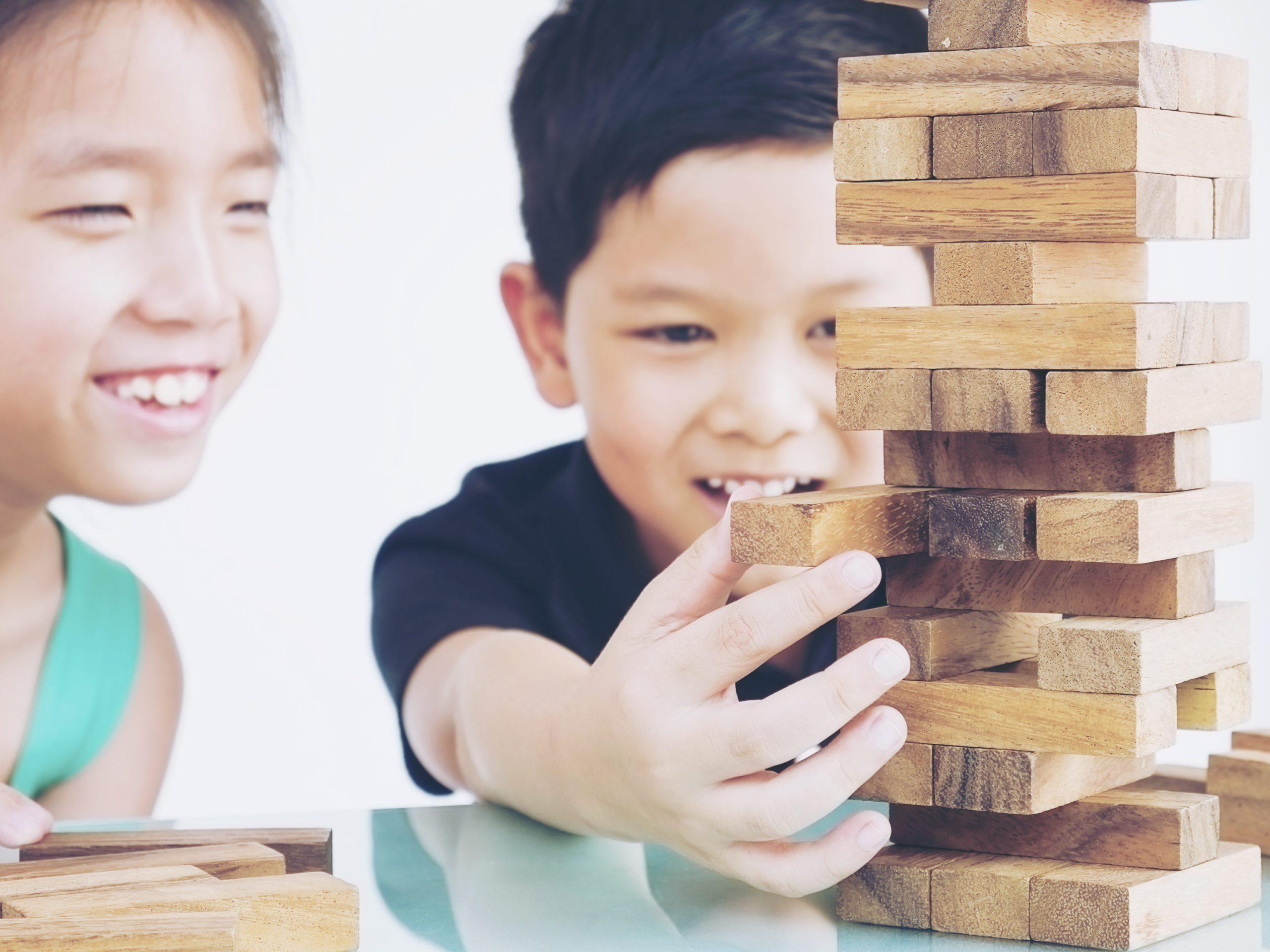 Two children playing a wooden block tower game, smiling and focused as one carefully pulls a block.