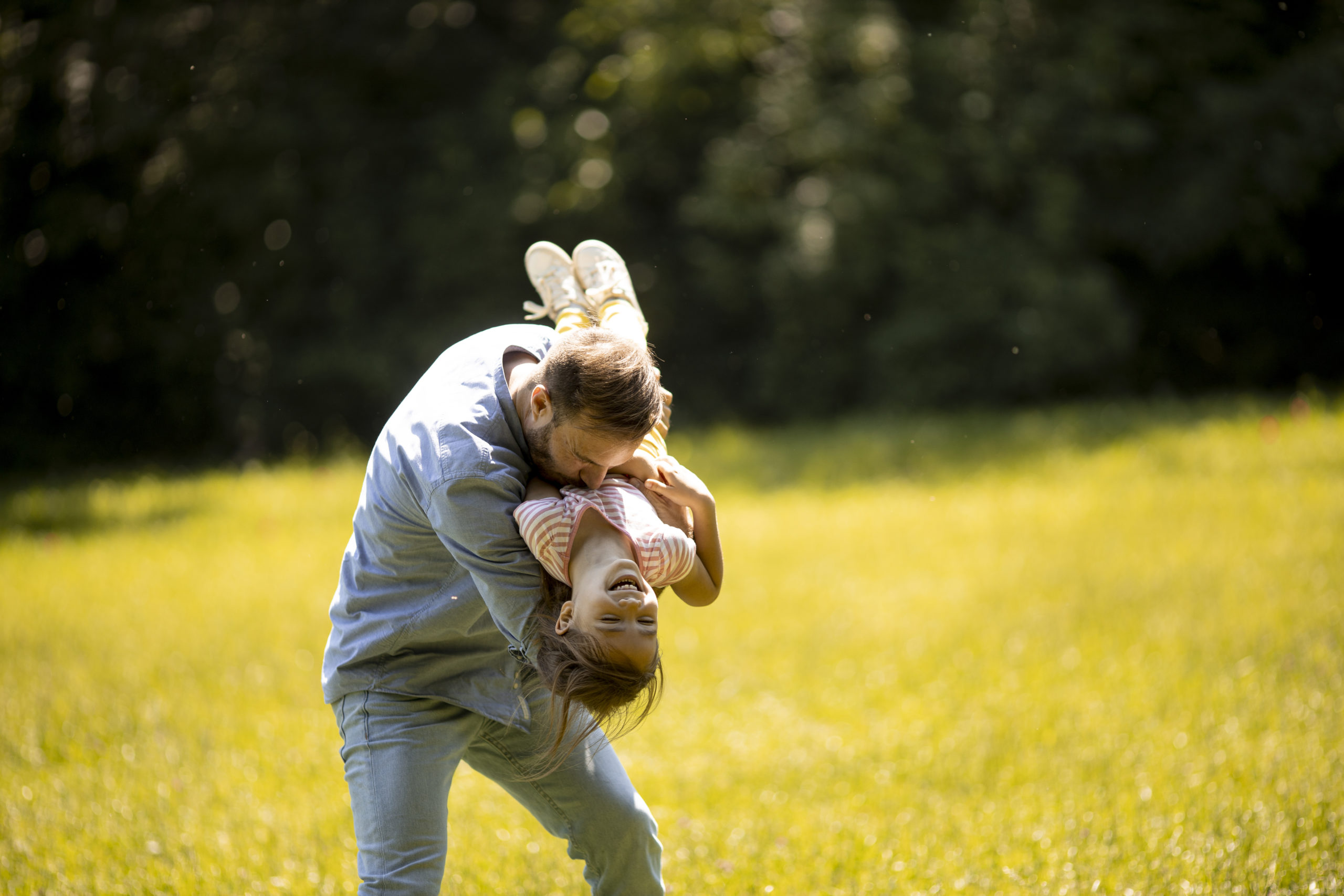 Father playing with his daughter outdoors in a sunny park, holding her upside down while they both laugh and enjoy a fun, carefree moment together.