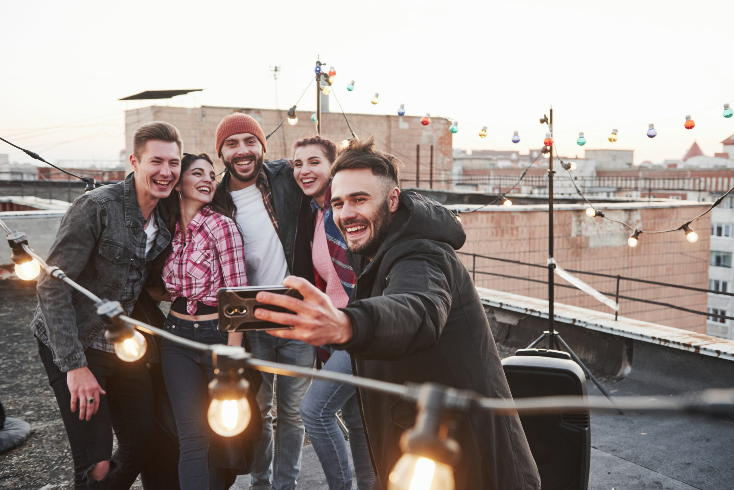 Group of young friends smiling and taking a selfie on a rooftop, surrounded by string lights and a cityscape in the background.