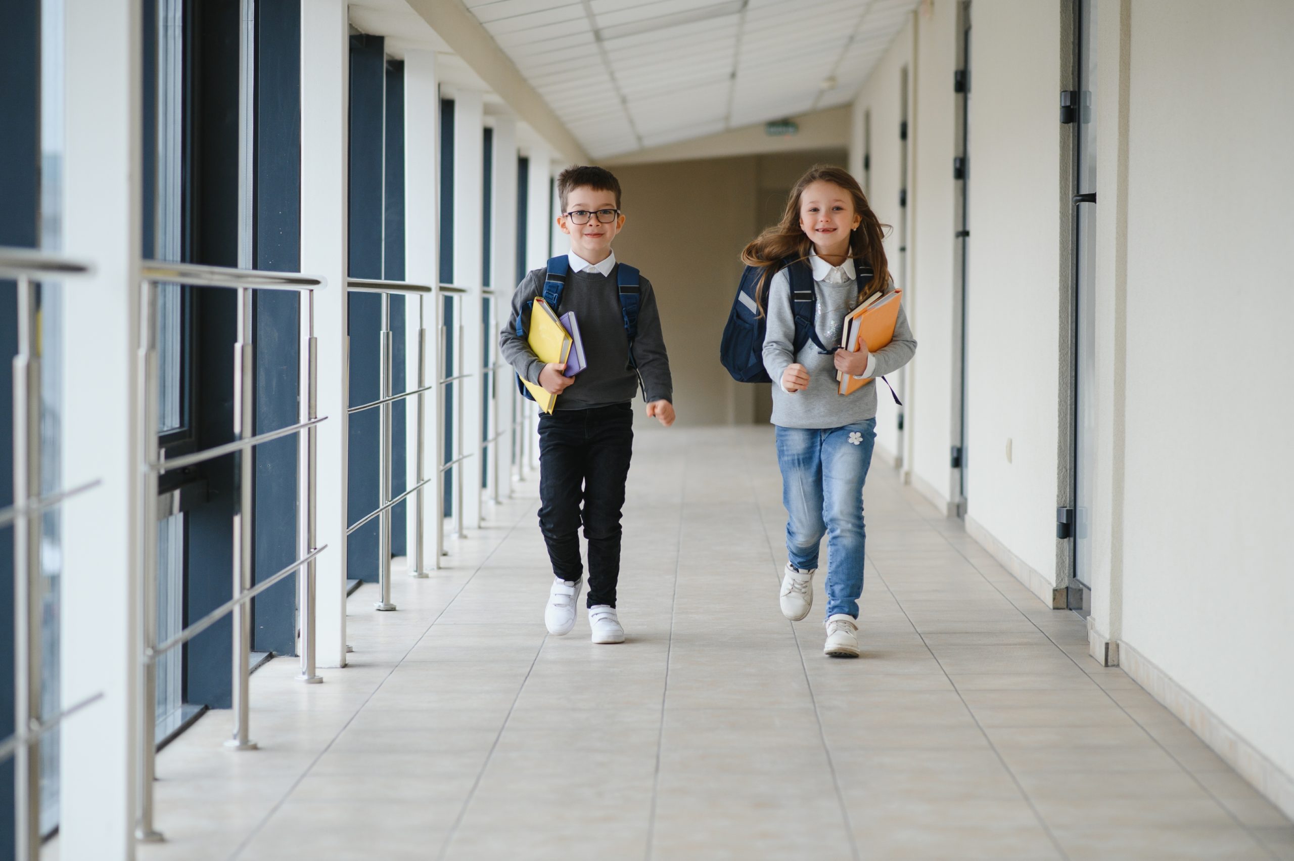 Two smiling schoolchildren with backpacks and books walking down a school corridor