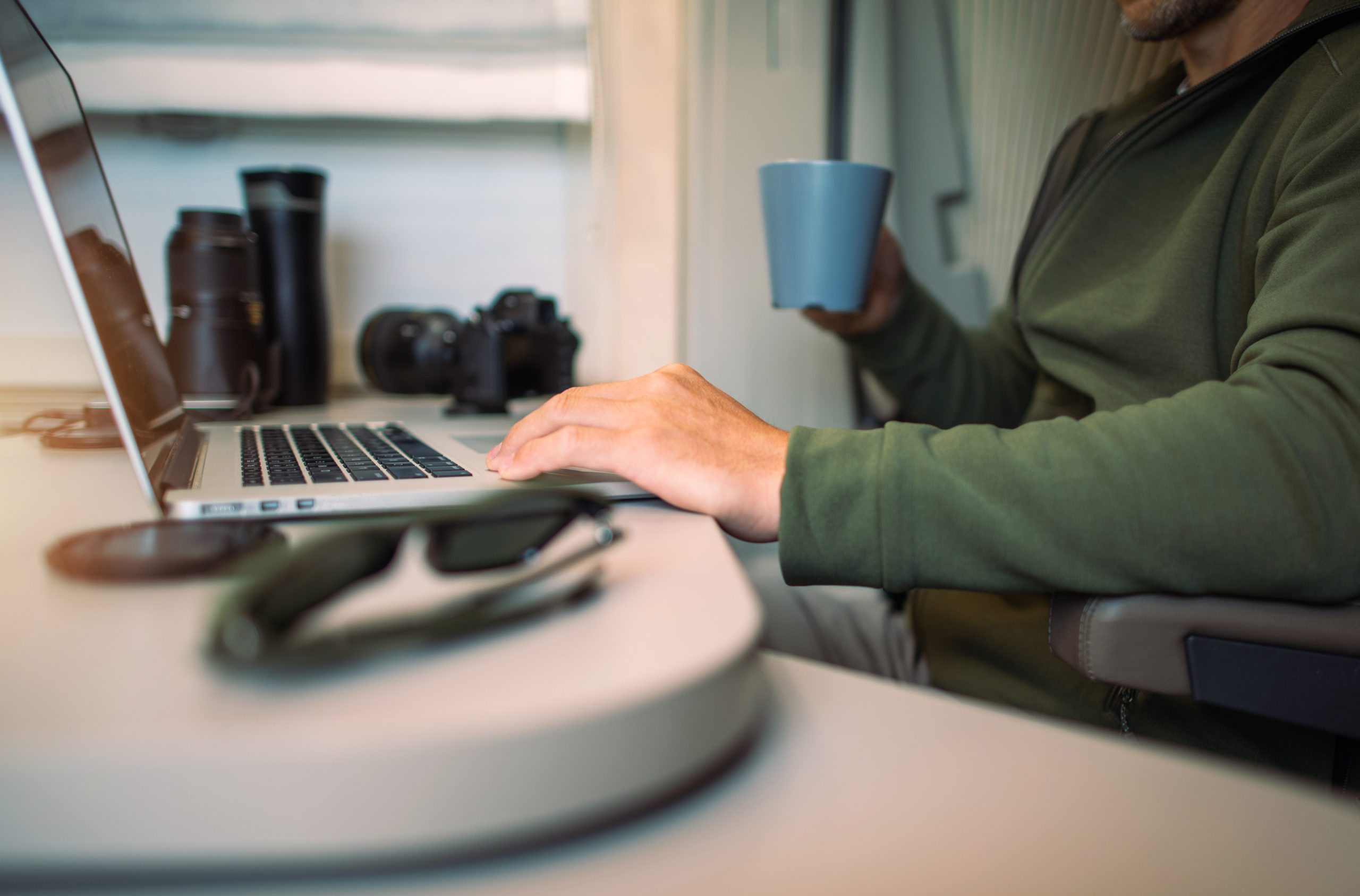 Person working on a laptop in a camper van, holding a mug, with camera equipment and sunglasses nearby.